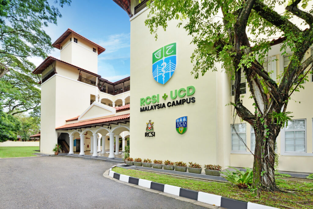 The exterior of the RCSI + UCD Malaysia Campus building. The building features a clean, modern design with white walls and red-tiled roofs. Prominent signage on the building displays the RCSI and UCD logos, along with the text 'RCSI + UCD Malaysia Campus.' The surrounding area includes lush green trees and well-maintained landscaping, creating a serene and welcoming environment.