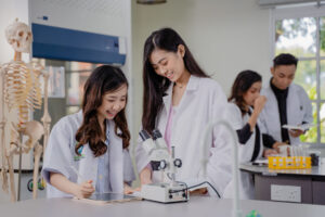 Two female medical students, wearing white lab coats, are smiling and engaged in a discussion while working with a microscope and a tablet in a laboratory setting. A skeleton model is visible in the background, along with other students working with lab equipment. The environment is bright and organized, indicating a collaborative and educational atmosphere.