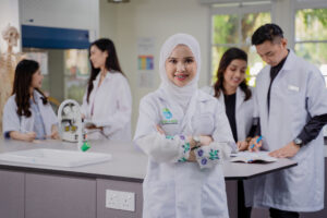 A group of medical students wearing white lab coats are gathered in a laboratory. In the foreground, a smiling female student in a hijab stands confidently with her arms crossed. In the background, other students are engaged in various activities, including reading a book and using lab equipment. The setting is bright and modern, with a skeleton model and scientific equipment visible.