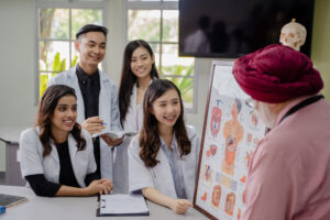 A group of medical students, dressed in white lab coats, are attentively listening to an instructor who is presenting an anatomical chart. The students are smiling and engaged, with one student holding a clipboard and another holding a book. The instructor, wearing a turban and pink shirt, is pointing at the chart. The setting is a bright, well-lit classroom with windows in the background.