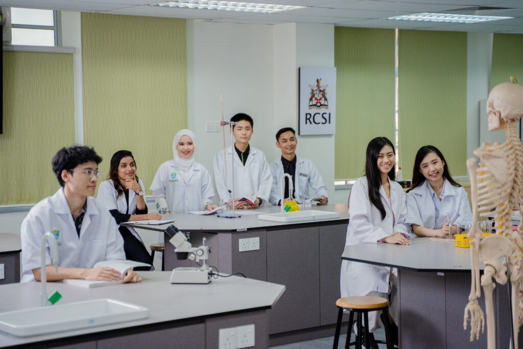 A group of medical students, wearing white lab coats, are attentively listening to a lecture in a modern laboratory classroom. They are seated and standing around lab tables equipped with scientific equipment, including microscopes and a skeletal model. The students are engaged and smiling, indicating an interactive and positive learning environment. The classroom has large windows and RCSI signage on the wall, adding to the professional academic setting.