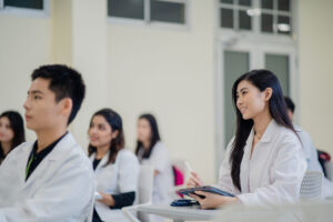 Medical students are attentively listening in a classroom setting. They are dressed in white lab coats and seated at desks with notebooks and tablets. The focus is on a female student smiling and engaged, while other students are slightly blurred in the background. The classroom environment is bright and modern, fostering a conducive learning atmosphere.