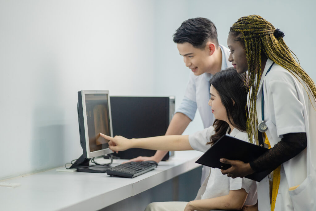 Three medical students, one male and two females, are gathered around a computer screen in a clinical setting. One student is seated and pointing at the screen while holding a tablet, while the other two students stand close, observing and discussing the displayed image. The students are dressed in white lab coats, and the environment appears to be a modern and clean laboratory or classroom.