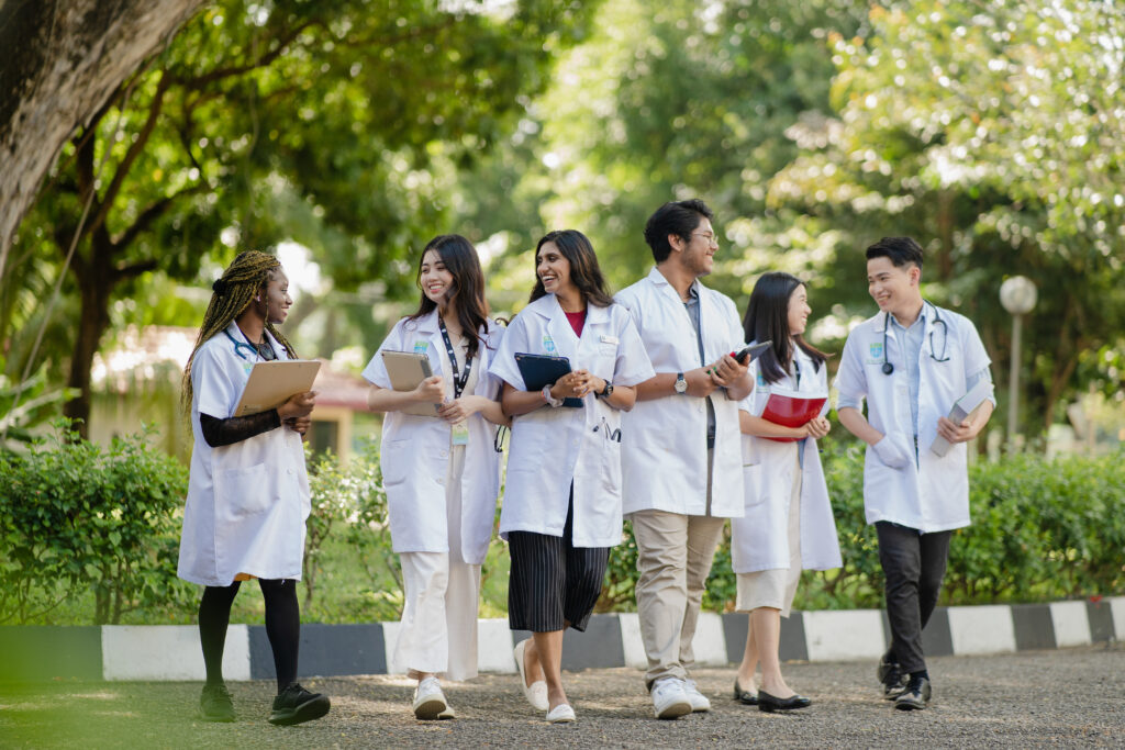 Six medical students, wearing white lab coats, are walking together outdoors in a lush, green campus setting. They are engaged in conversation and smiling, each carrying books, tablets, or clipboards. The environment is bright and welcoming, indicating a collaborative and friendly atmosphere among the students.